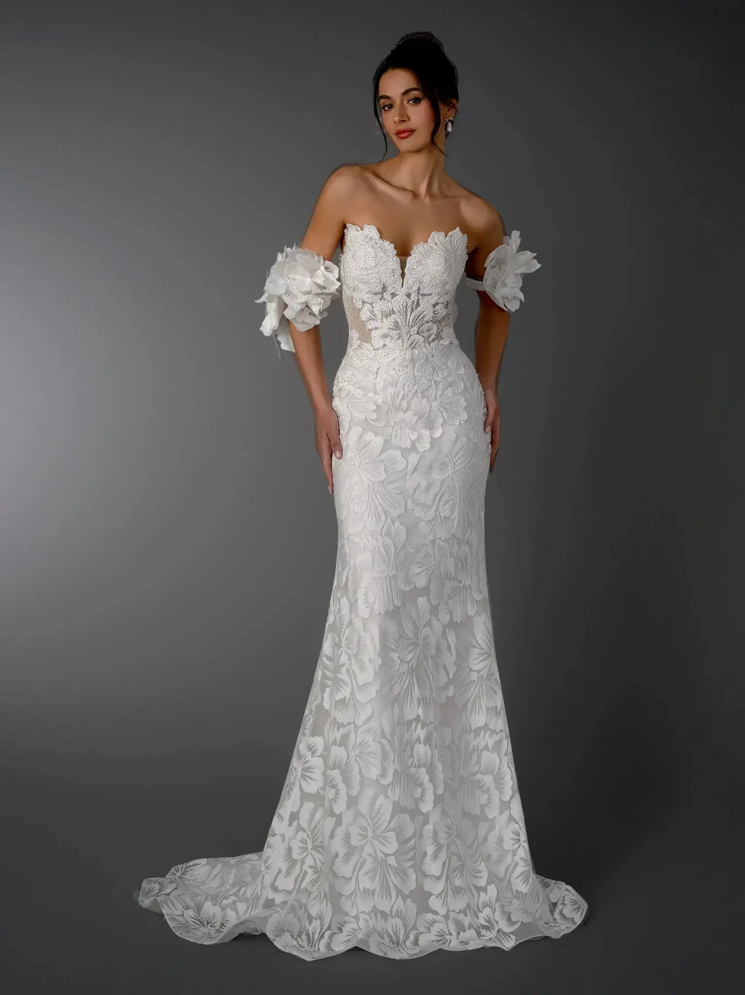 A woman wearing an elegant white wedding dress with floral patterns and off-the-shoulder sleeves, standing against a dark background.