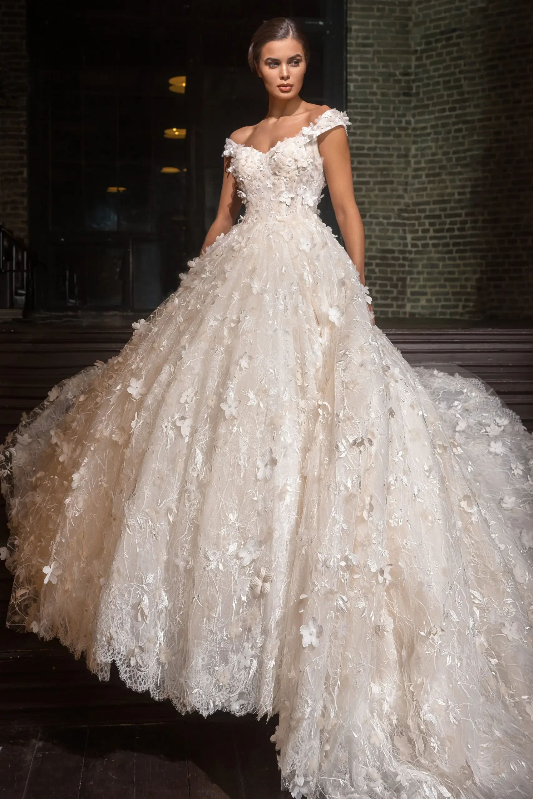 Model is wearing an off-shoulder ball gown with lace and tulle layers, Austrian crystal beading, and a full, dramatic skirt.