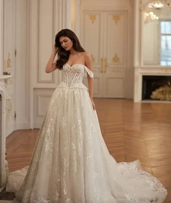 A woman wearing an elegant, off-the-shoulder wedding dress with intricate detailing, standing in a beautifully decorated interior space.