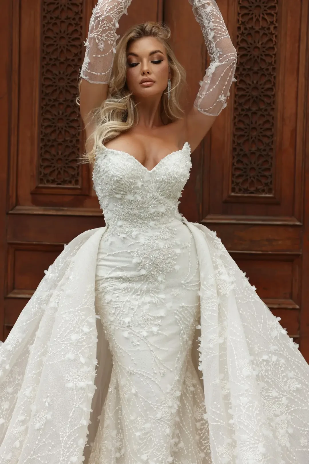 Model is wearing a pointed cat-eye bodice gown with 3D embellishments and a layered voluminous overskirt.