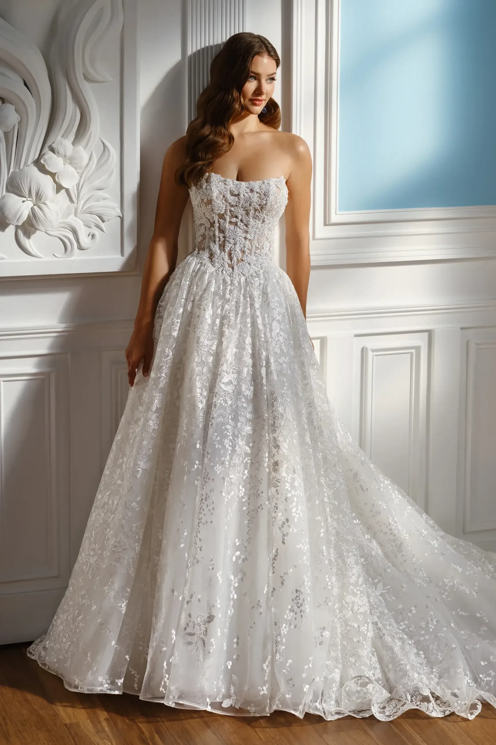 A woman in a strapless, embellished white wedding dress stands against a decorative wall, with soft lighting illuminating the scene.