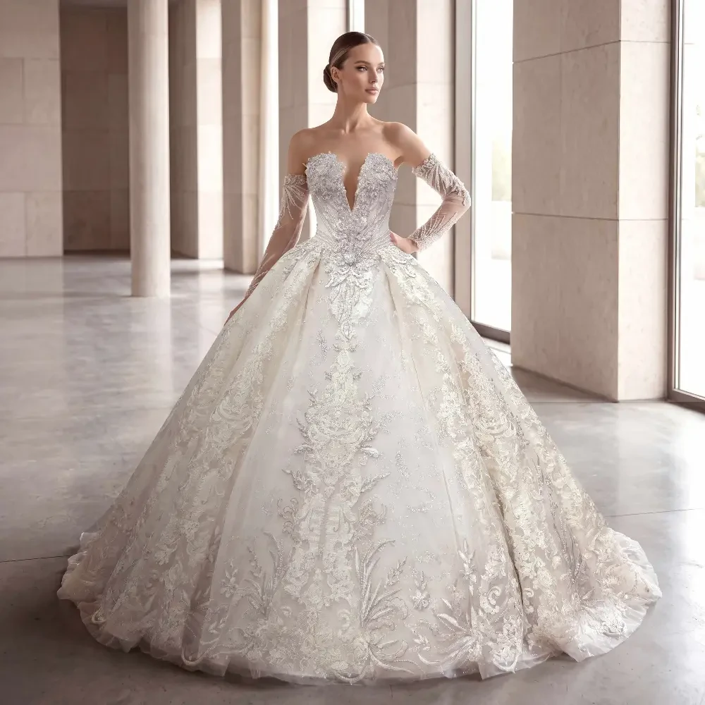 A woman in a stunning off-the-shoulder wedding gown with intricate lace and bead detailing, standing in a well-lit modern space.